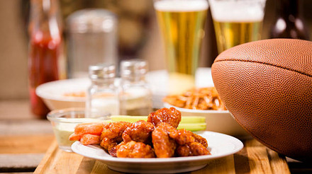 Sports bar, pub setting. Spicy chicken wings foreground. Football, pretzels, peanuts, beer on bar counter top. Dartboard in background.